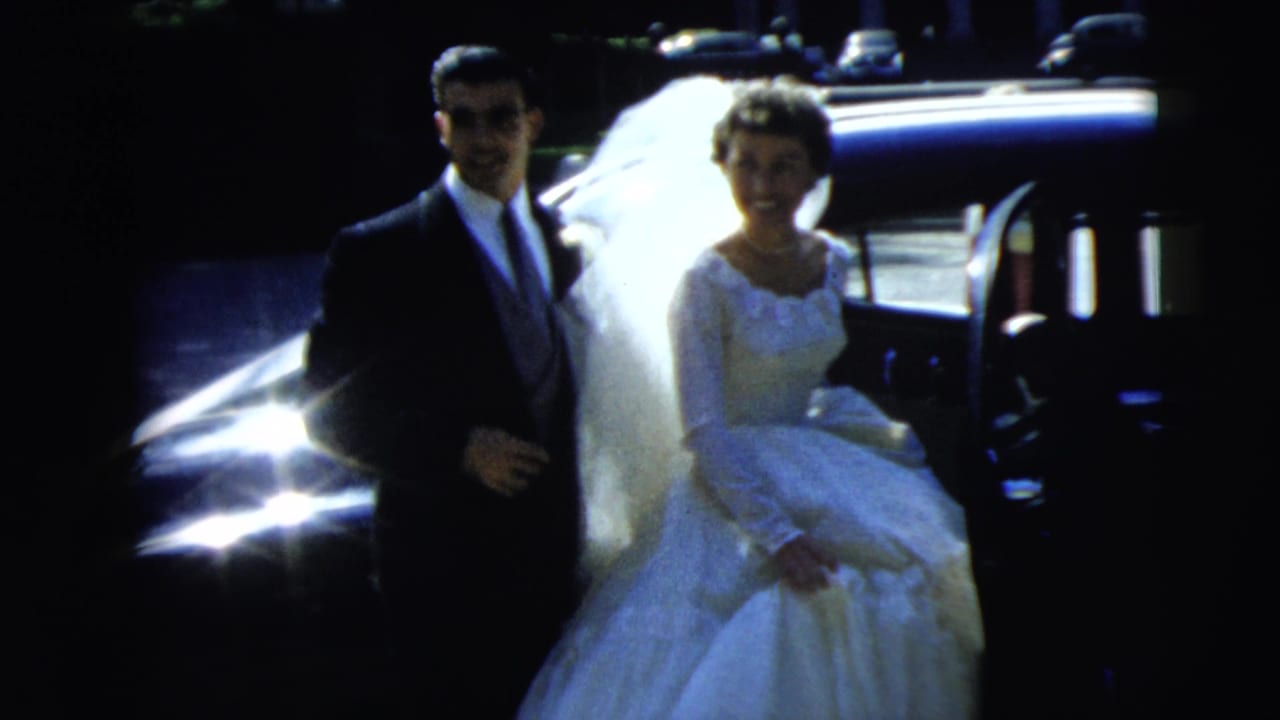 1962 Camden wedding home-movie frame showing a bride in a white gown and veil with a groom in a dark suit beside a car.