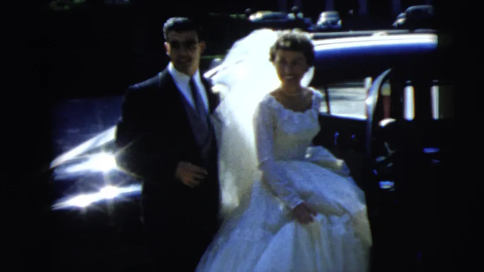 1962 Camden wedding home-movie frame showing a bride in a white gown and veil with a groom in a dark suit beside a car.