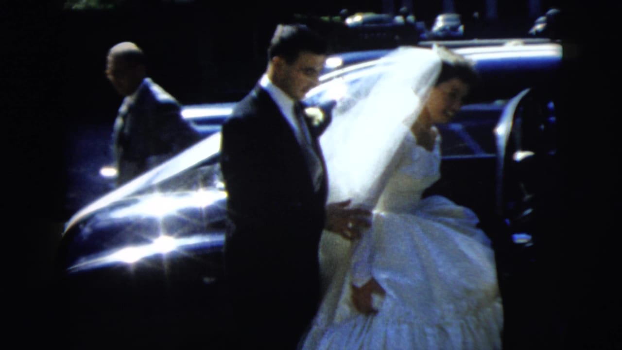 Groom helps the bride near an open car door while her veil stretches across the frame.