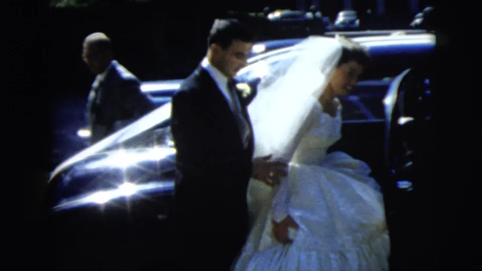 Bride and groom at a Camden, New Jersey wedding in 1962, standing beside a dark car as she gathers her gown.