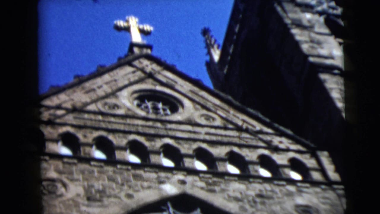 1962 8mm frame of a Camden stone church roofline, showing a gold cross, triangular gable, and arched openings against blue sky.