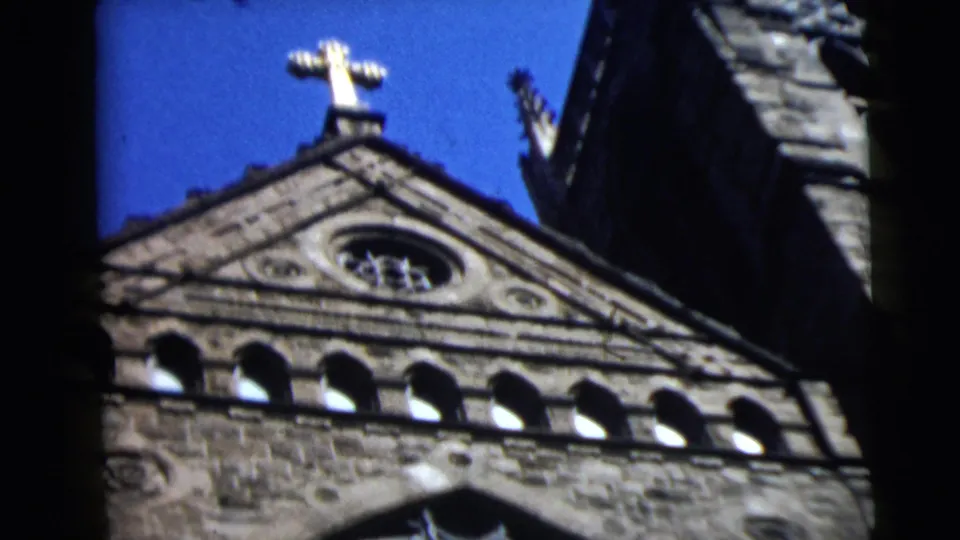 1962 8mm frame of a Camden stone church roofline, showing a gold cross, triangular gable, and arched openings against blue sky.