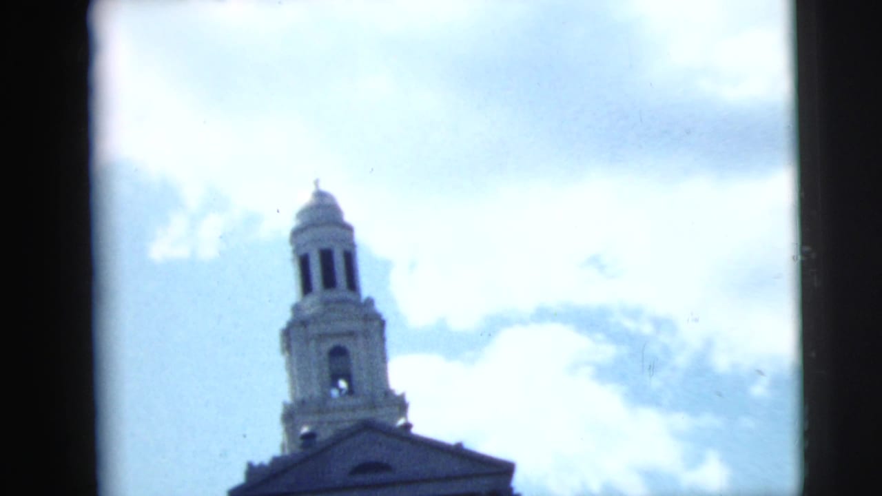 1962 Camden, New Jersey 8mm frame looking up at a church bell tower and roofline against bright clouds.