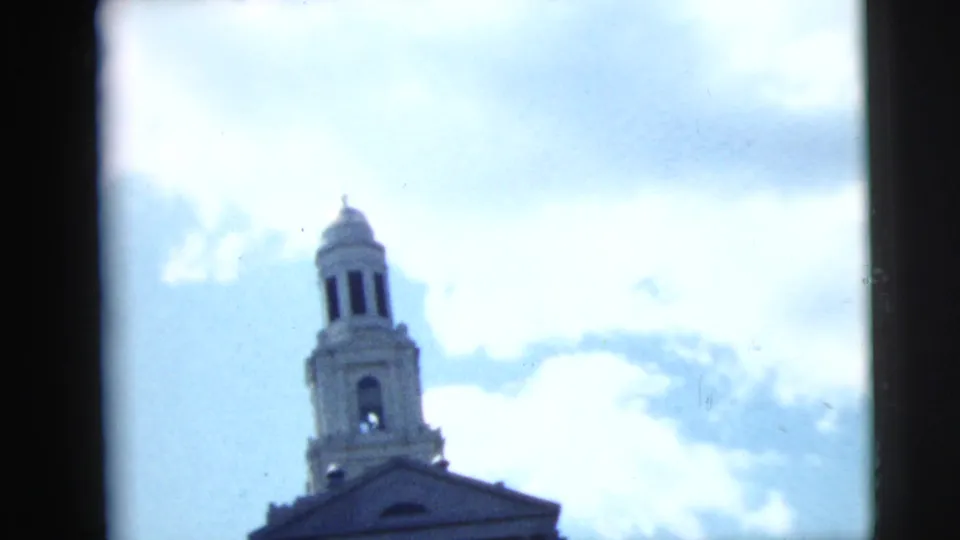 1962 Camden, New Jersey 8mm frame looking up at a church bell tower and roofline against bright clouds.