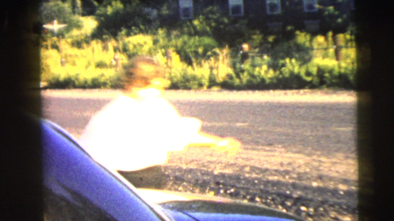 Adult woman in a white blouse and dark skirt appears beside a dark car on a sunny Camden street in the 1962 home movie.