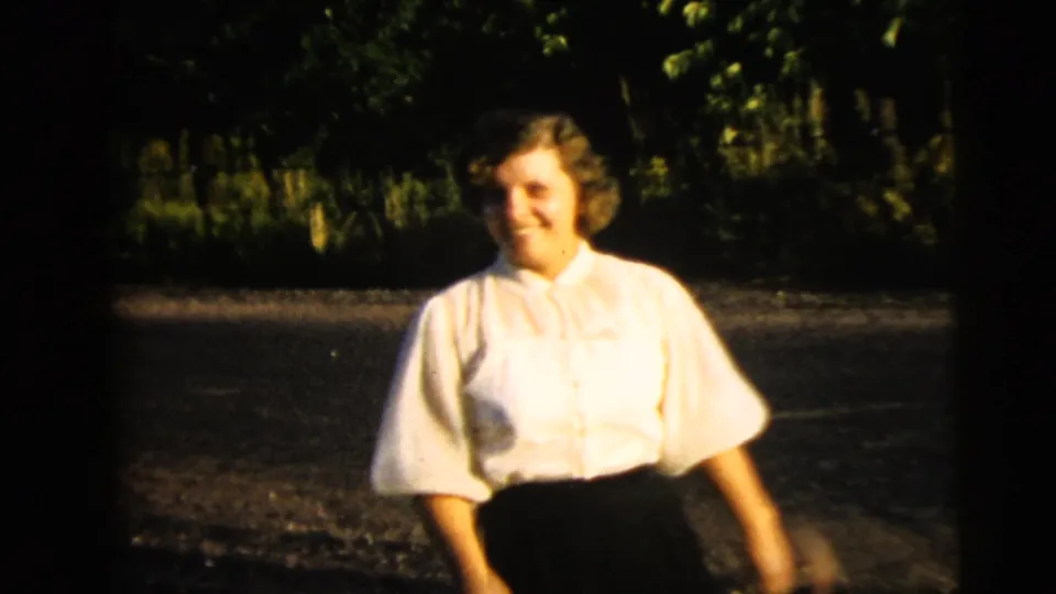 The woman walks past the camera in a white blouse and dark skirt, with trees and pavement behind her in the 1962 Camden footage.