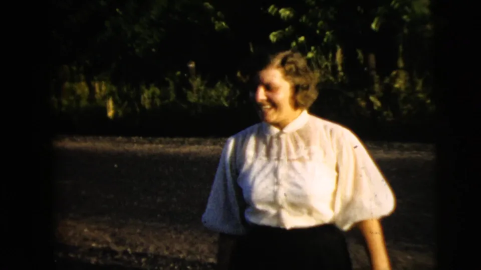 Smiling adult woman in a white blouse and dark skirt faces to the side on a shaded Camden street in the 1962 home movie.