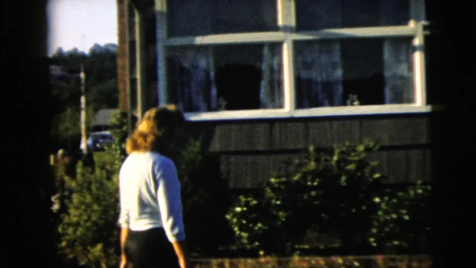 The woman is seen from behind near shrubs and a windowed house exterior in the Camden backyard.