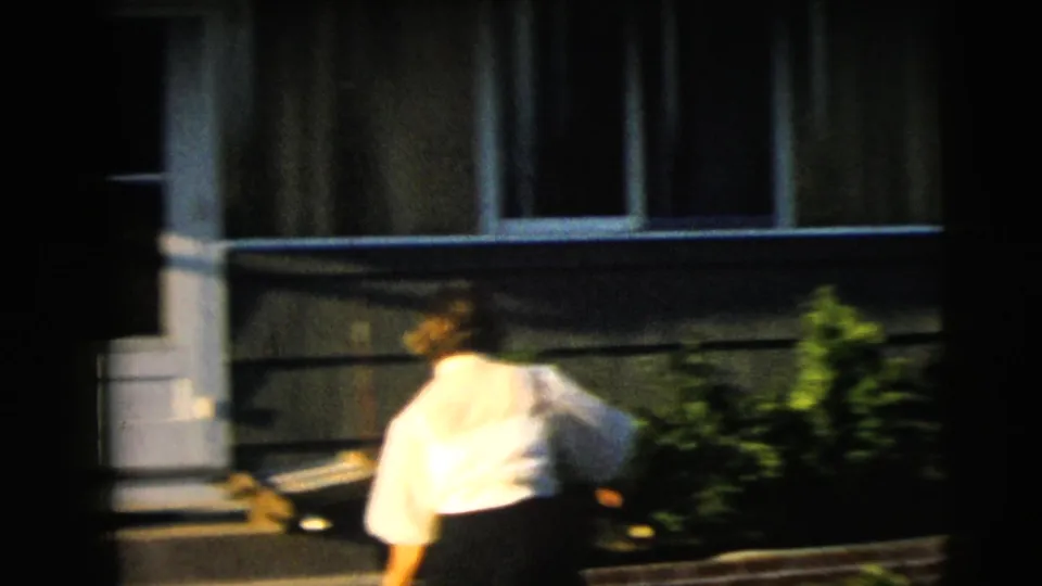 The woman moves near the house steps and shrubs, captured with motion blur on 8mm film.