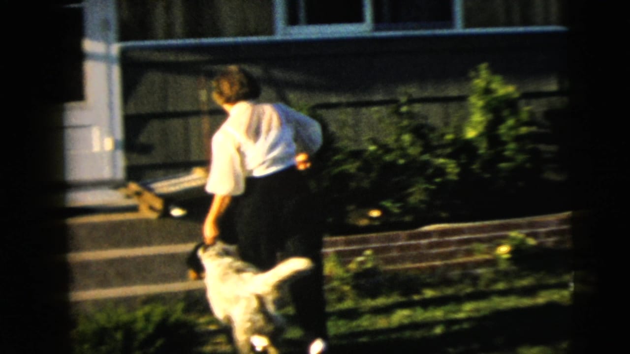 1962 Camden home-movie frame showing a woman in a white blouse and dark pants walking a large light-colored dog near a suburban front step.