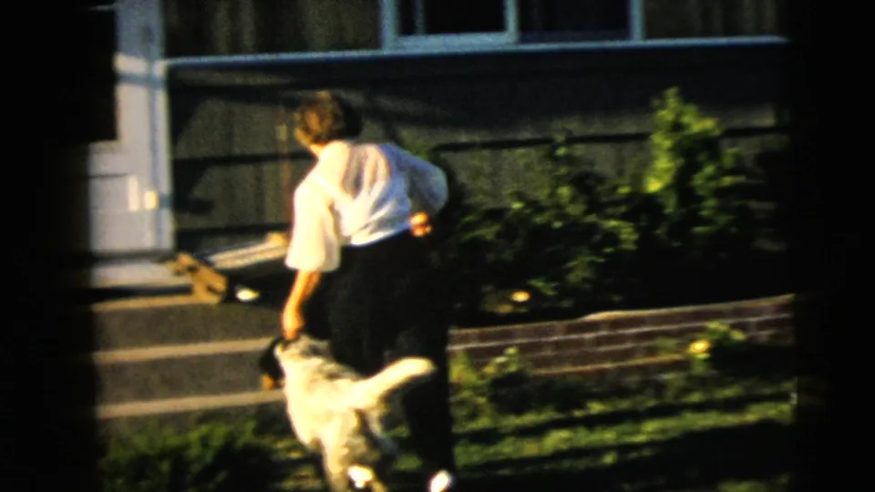 1962 Camden home-movie frame showing a woman in a white blouse and dark pants walking a large light-colored dog near a suburban front step.