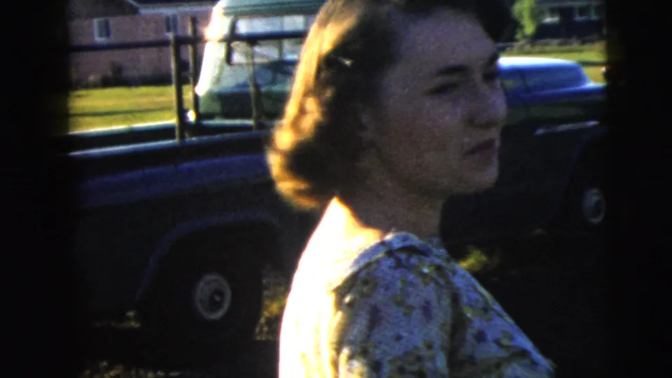1962 Camden home-movie frame showing a close view of a woman in a patterned top with a parked vehicle and lawn behind her.