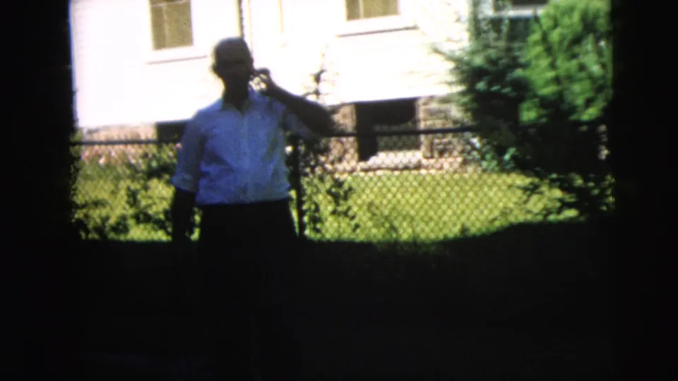1962 Camden exterior frame showing a man in a light shirt standing near a fence and house while holding an old-style telephone receiver.