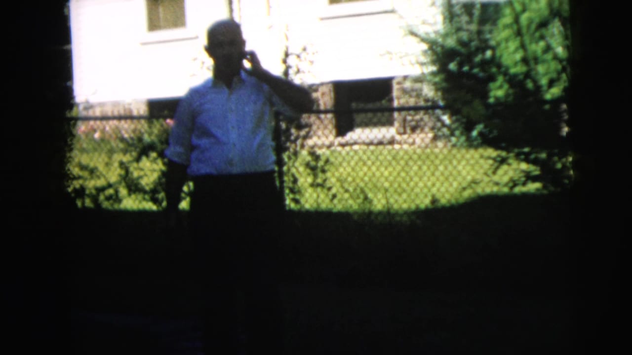 In 1962 Camden home-movie footage, a man stands by a lawn and chain-link fence outside a house while holding a phone to his ear.