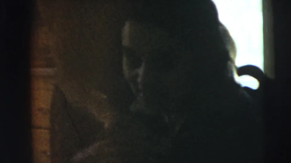 A dim interior home-movie frame shows a person in silhouette near a bright opening, with wall texture visible in low light.