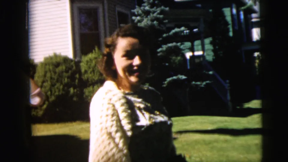 1962 Camden yard scene with a woman in a light sweater standing on grass near shrubs and house siding.