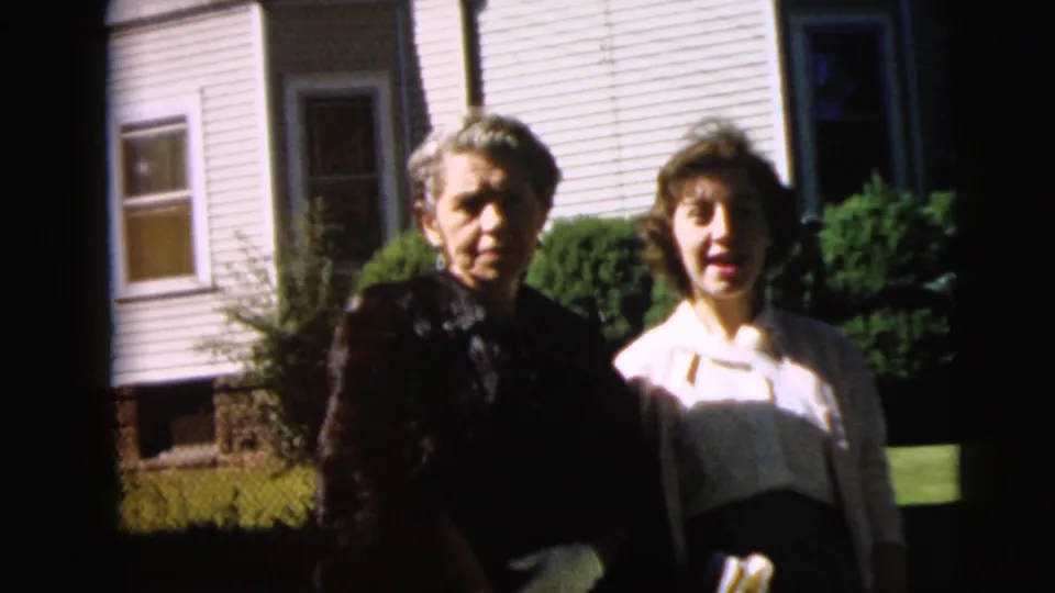 1962 Camden home-movie frame of two women outdoors near a white-sided residence, with a dark figure or hair in the foreground.