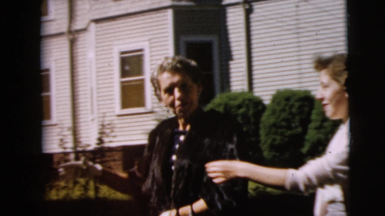 Two women stand outside a white clapboard house in bright sunlight.
