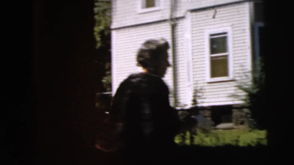 A dark vertical obstruction divides the frame as two people move near the residential exterior.