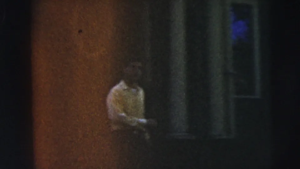 A dim handheld frame shows a person near a doorway or shaded wall during the Camden home-movie scene.