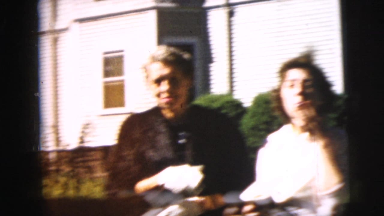 Two young adults seated or standing near a white house in a sunlit yard.