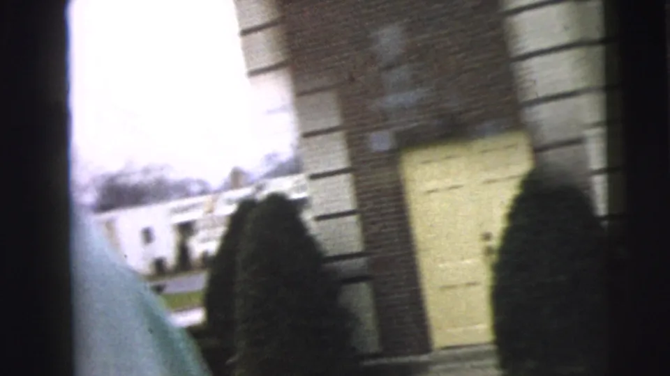 The handheld Camden footage briefly frames a brick residential building with a pale door, shrubs, lawn, and the edge of a coat in the foreground.