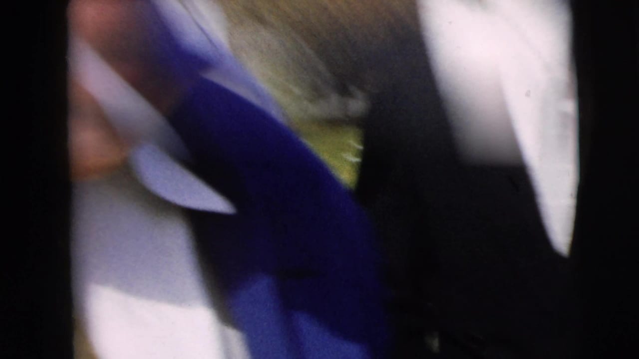 Tight, handheld 8mm close-up of a person's face and shoulder, with another figure or coat passing close to the lens.