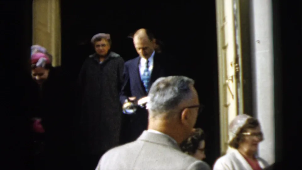 People in long coats and formal clothing exit a doorway in Camden, New Jersey, including a man in a suit and tie among the group.