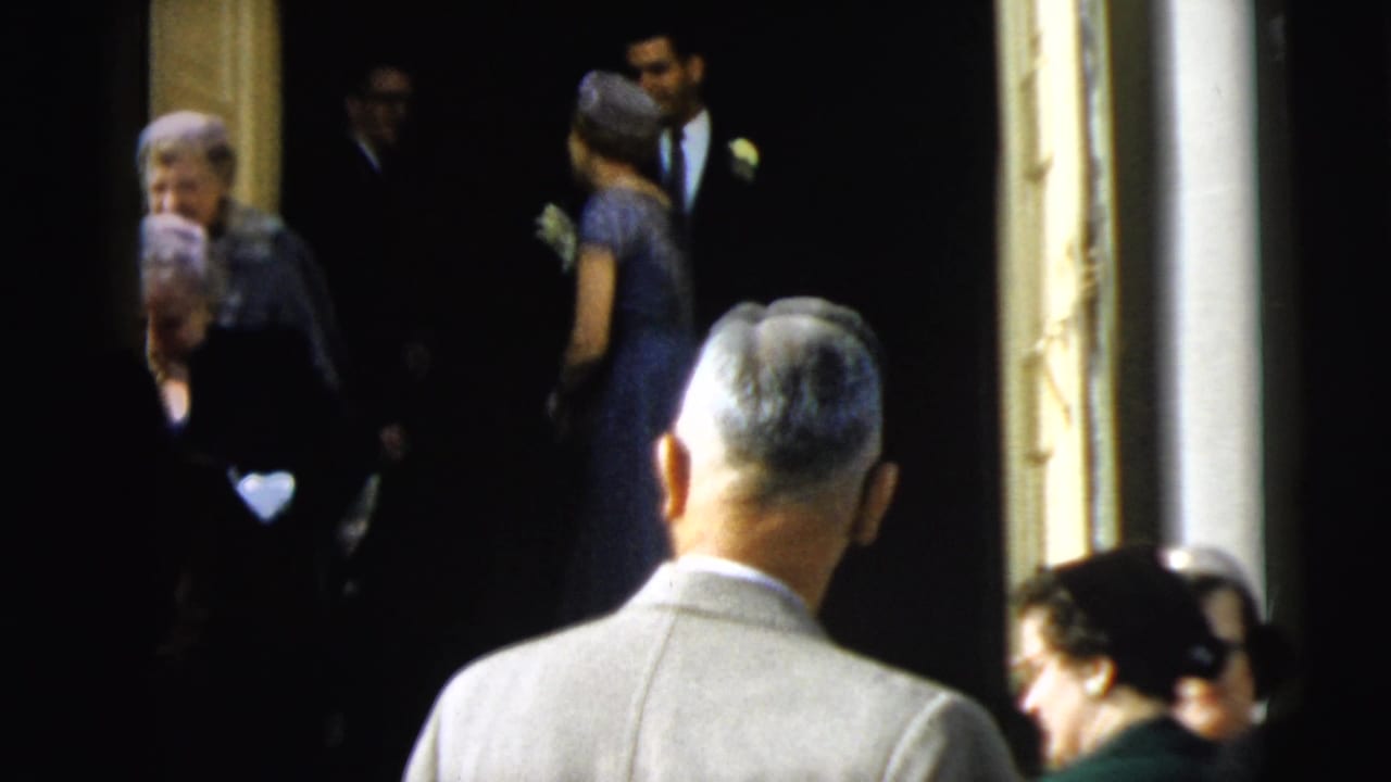 A formal group, including women in hats and a suited man, appears at a Camden doorway during the 1962 home-movie sequence.