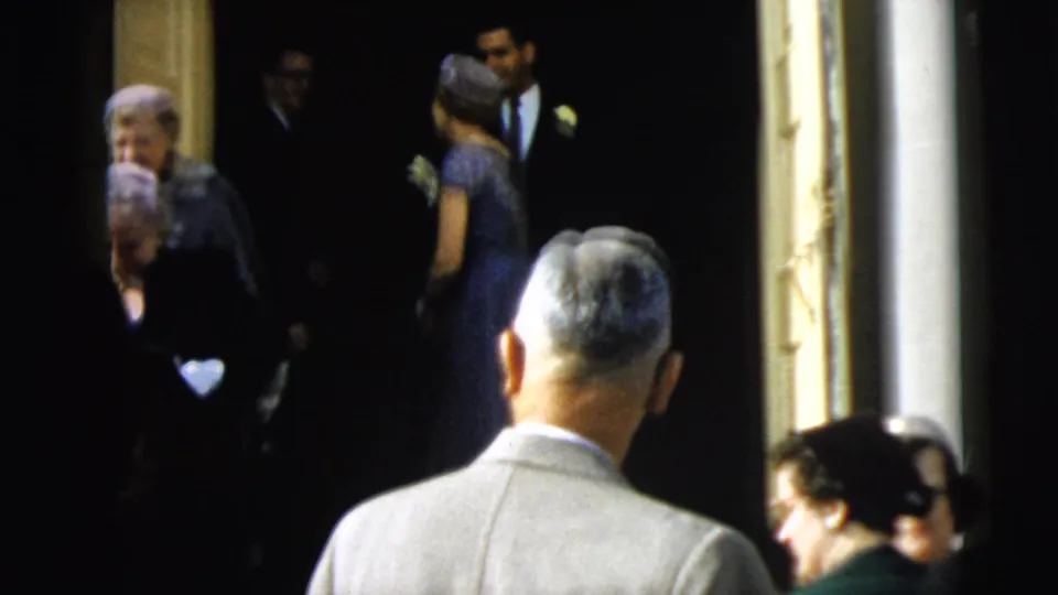 A formal group, including women in hats and a suited man, appears at a Camden doorway during the 1962 home-movie sequence.