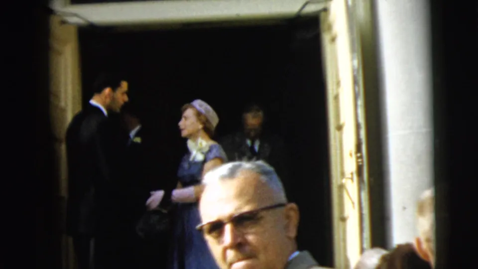 A woman in a blue dress and hat stands near a suited man at the Camden building entrance, partially obscured by a close foreground passerby.