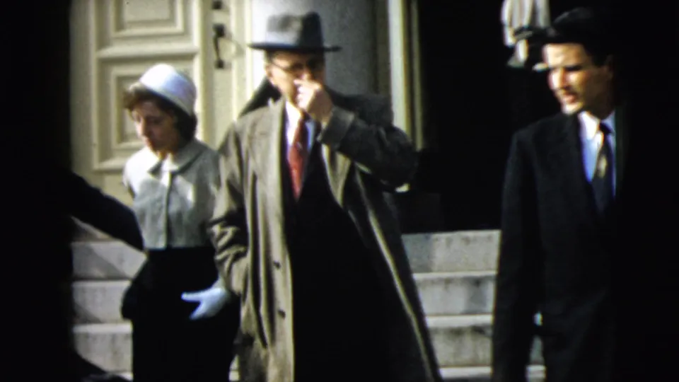 At the Camden building exit, a man in a trench coat and fedora descends steps as other formally dressed adults move nearby.