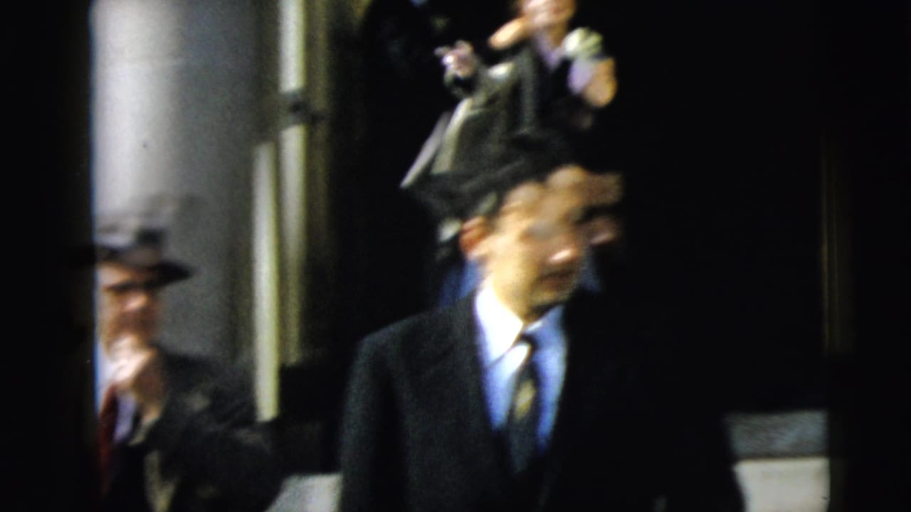 Man in a suit and brimmed hat walks past a stone entrance with other formally dressed guests nearby.