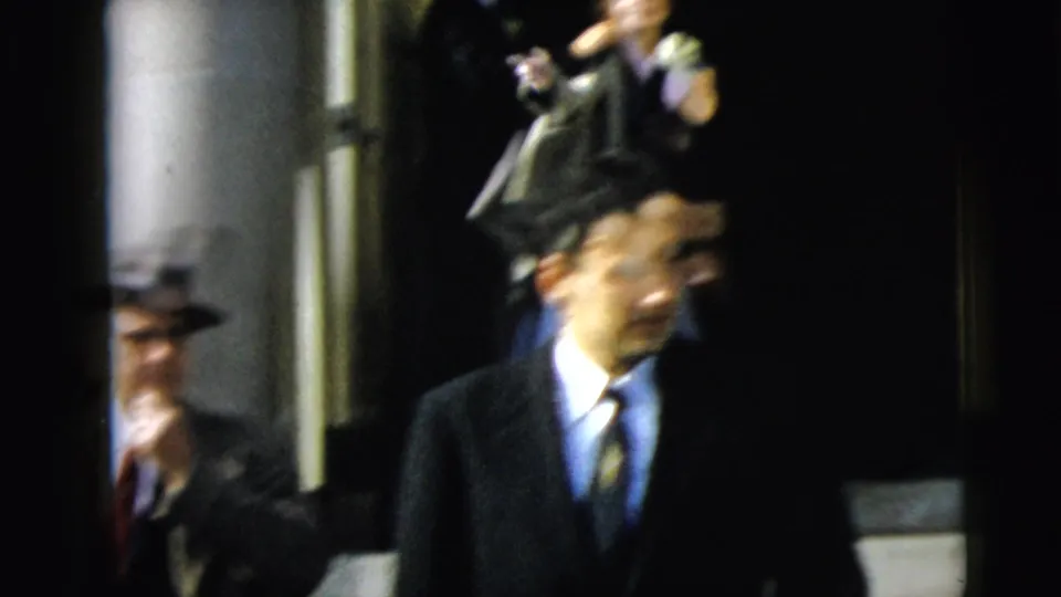 1962 Camden home-movie frame showing a suited man in a brimmed hat passing a stone building entrance during a formal gathering.