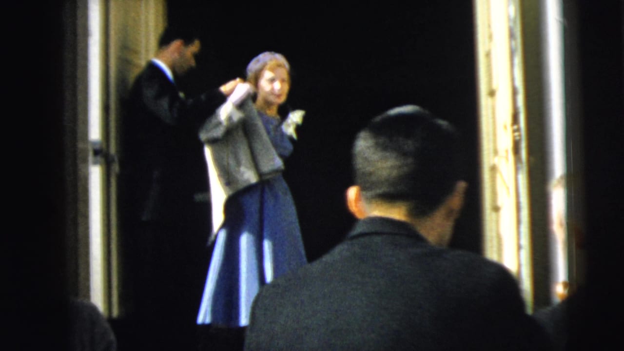 Woman in a blue dress and light hat adjusts her jacket near open doors while a suited man stands nearby.