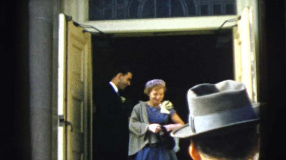 1962 Camden home-movie frame with a woman in blue at an open doorway and a foreground brimmed hat crossing the view.