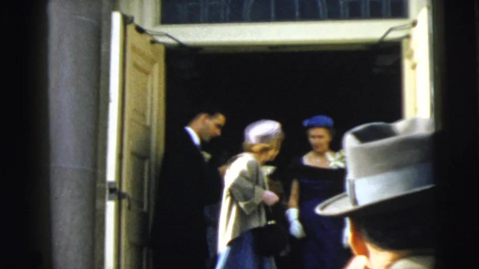 1962 Camden frame showing formally dressed women and suited men gathered at an open stone building entrance.