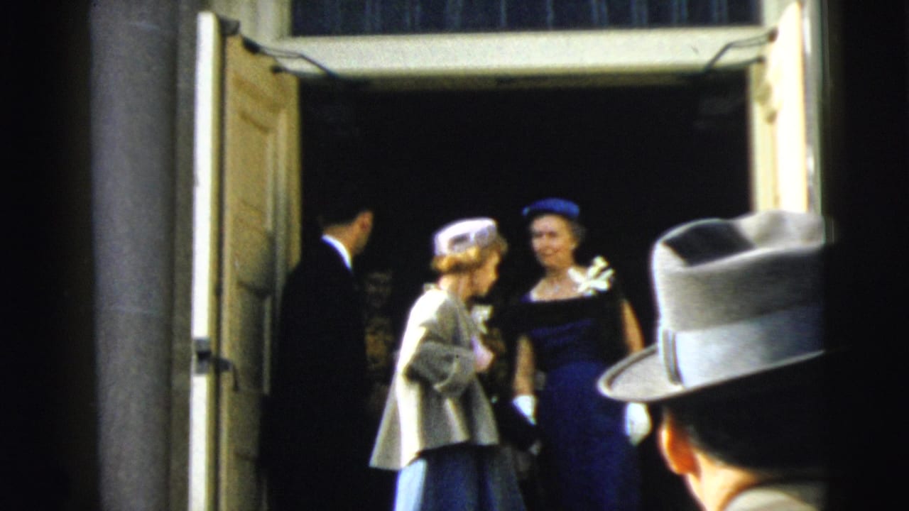 In 1962 Camden home-movie footage, wedding guests in hats and formal clothing stand near open doors as the camera looks out from inside.