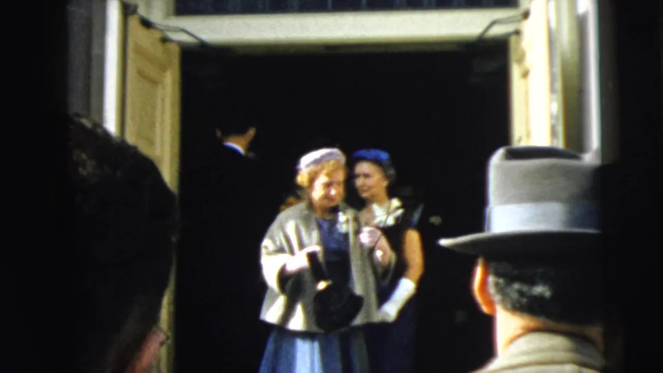 Two women in 1960s formal wedding attire pause in the open doorway, with a man in a suit partly seen behind them.