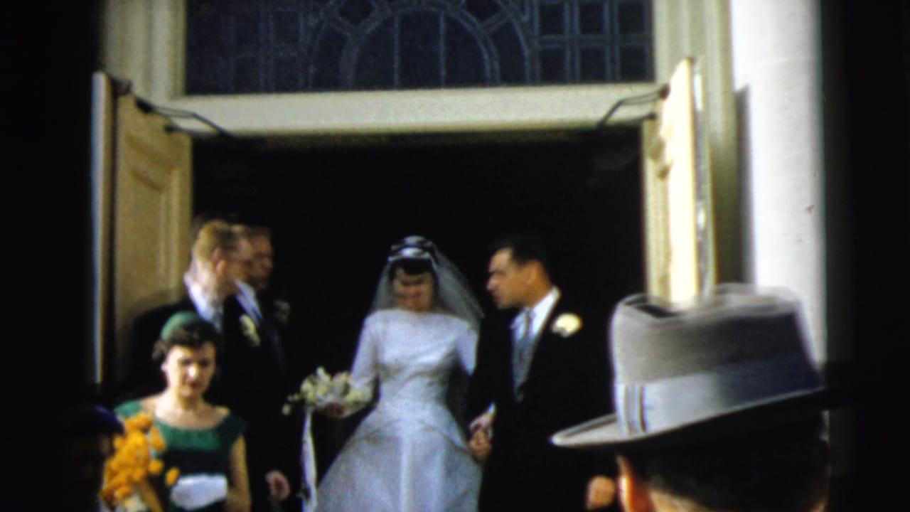Bride and groom walk arm in arm from the church with attendants behind them.
