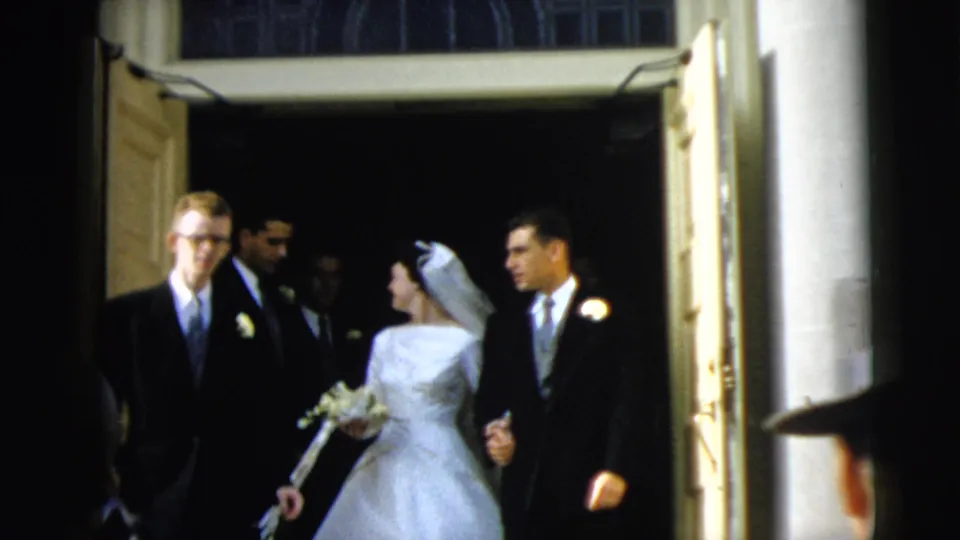 At the Camden church exit, the bride looks toward the groom while a suited guest moves across the foreground.