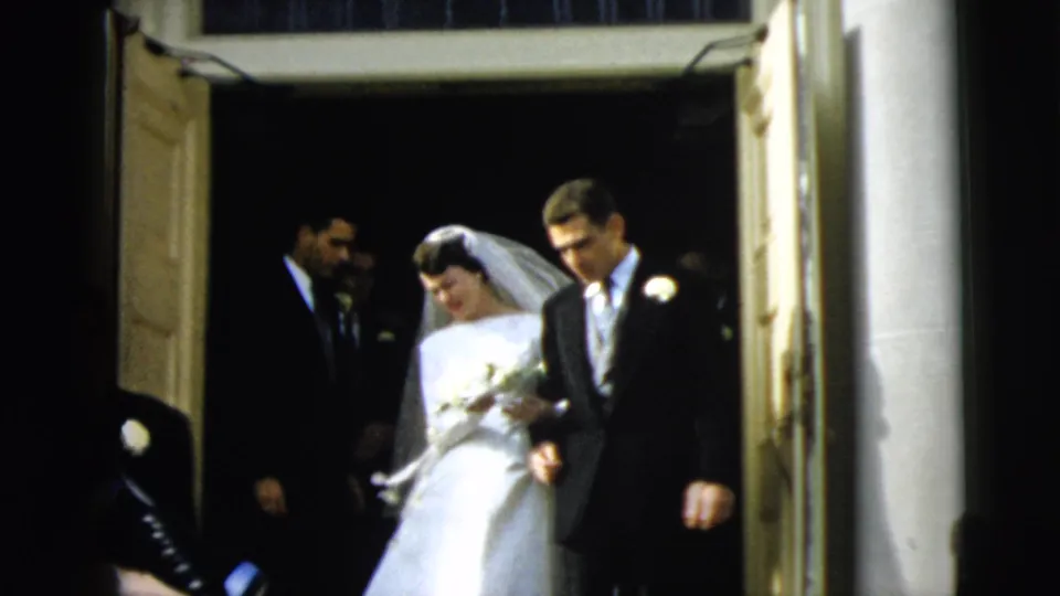 The bride and groom continue out of the Camden church entrance, framed by open doors, columns and attendants behind them.