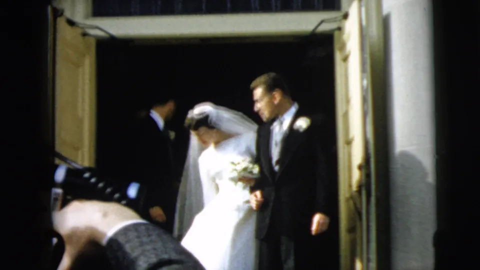 In Camden, New Jersey, 1962, a bride in a white gown and veil exits a church doorway with the groom beside her.