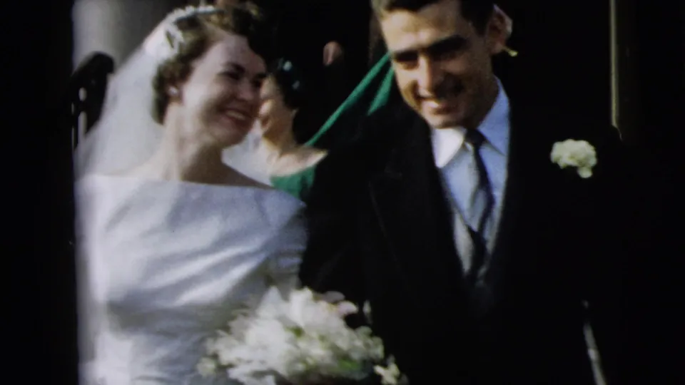 In 1962 Camden wedding footage, a bride with bouquet and groom in dark suit smile while walking past guests.