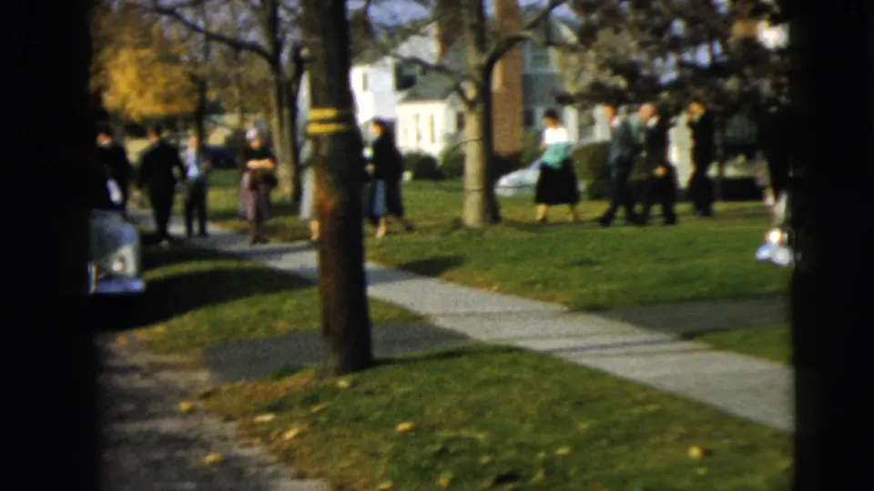 A Camden residential street scene shows wedding guests walking on a sidewalk among lawns, trees, houses, and parked cars.