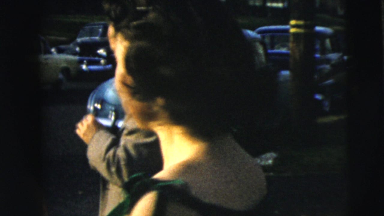 In 1962 Camden home-movie footage, a woman in a dark dress with green trim turns near parked cars on a residential street.