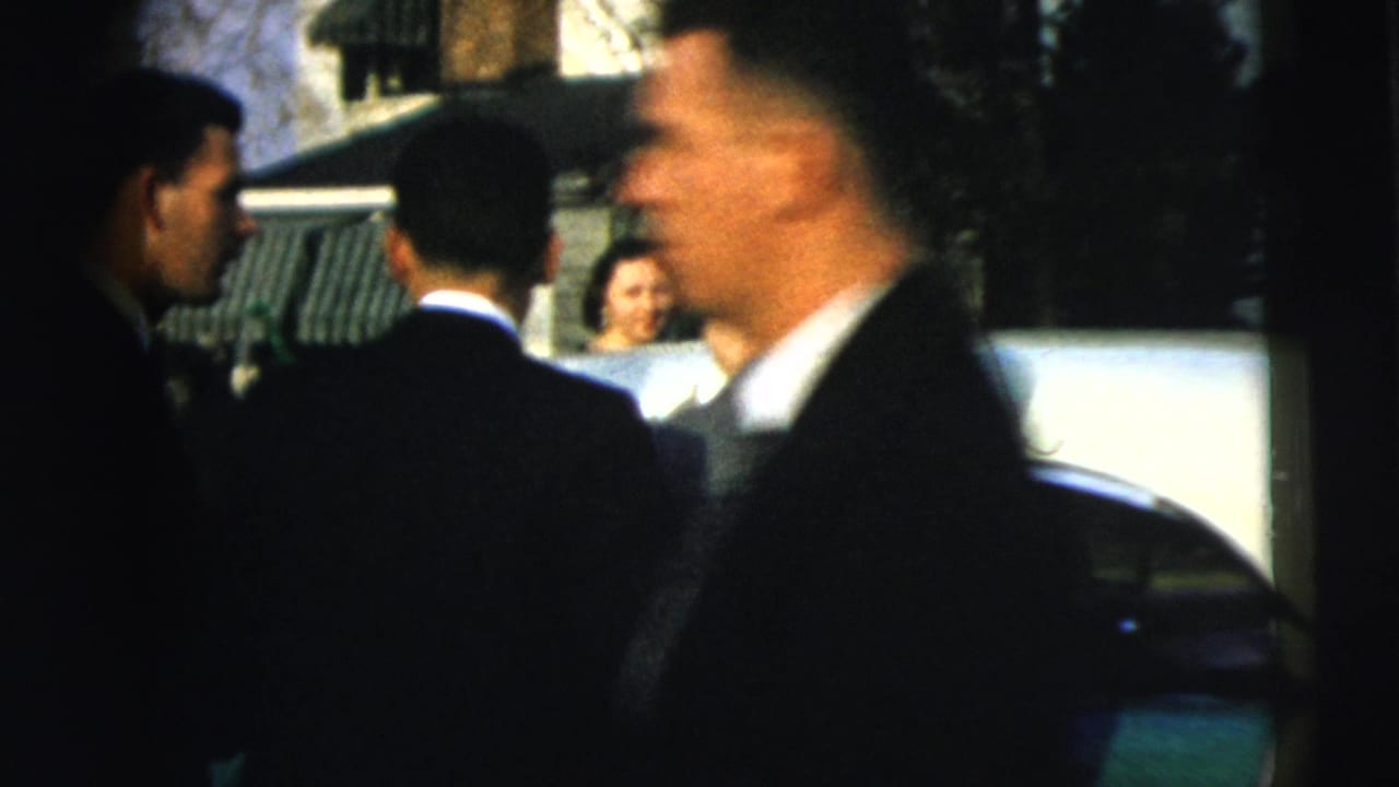 Suited men stand beside a car during a 1962 Camden wedding departure, with guests and houses visible behind them.
