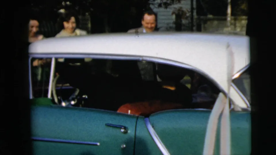 A two-tone green and white car fills the frame while wedding guests smile behind it in the 1962 Camden home movie.