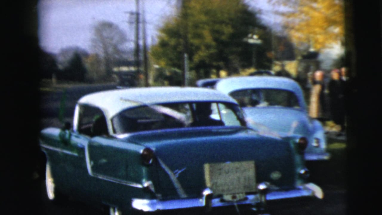 A green-and-white sedan drives ahead on a residential Camden street, with another pale car visible behind it.