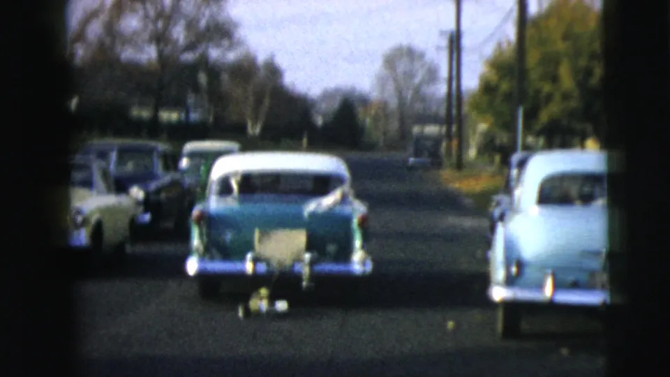 The green-and-white sedan travels among other mid-century cars on a Camden residential road, with trees and utility poles in view.